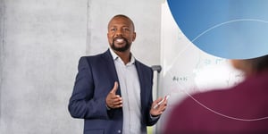 man standing next to white board and speaking to a group of people