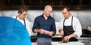 man holding tablet standing between one male cook and a female cook talking to male cook 